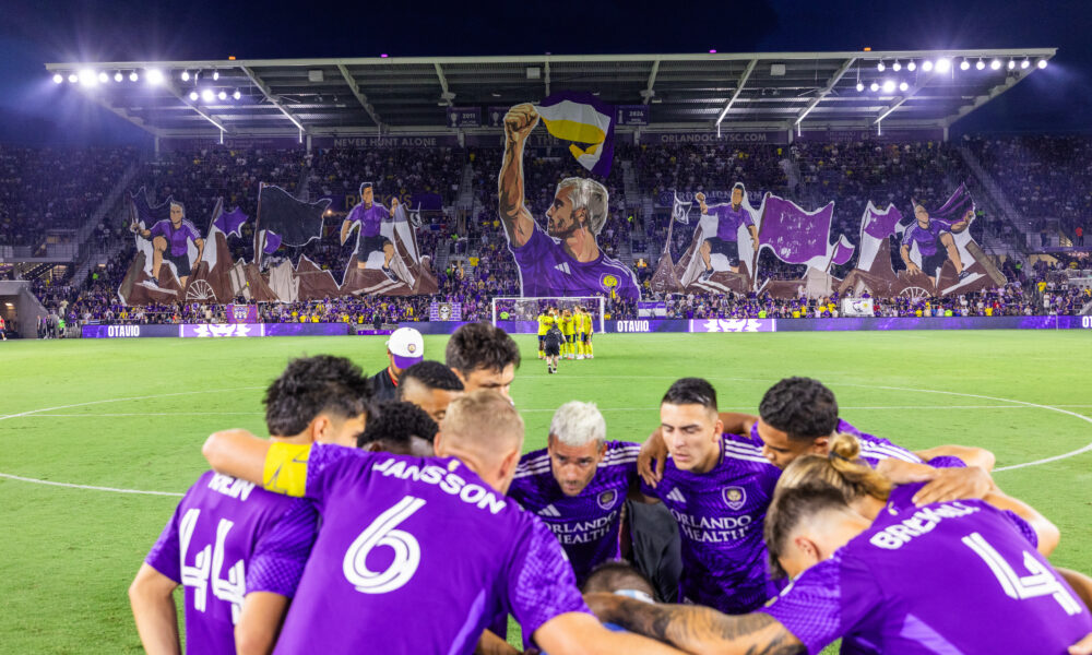Image of Orlando City players huddled together with a tifo displayed in the background.